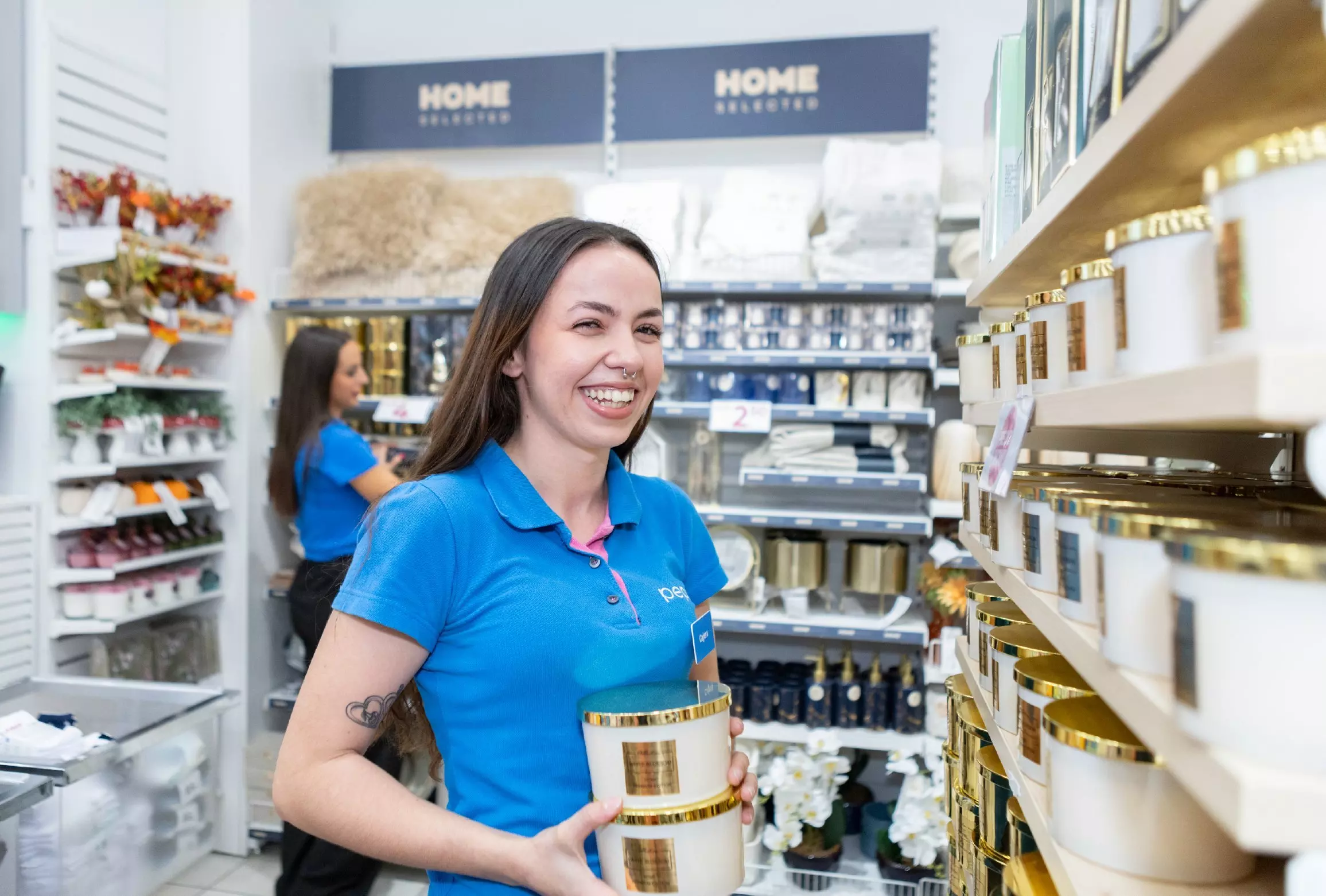 Angajată zâmbitoare în tricou polo albastru ținând o lumânare decorativă într-un magazin de produse pentru casă.