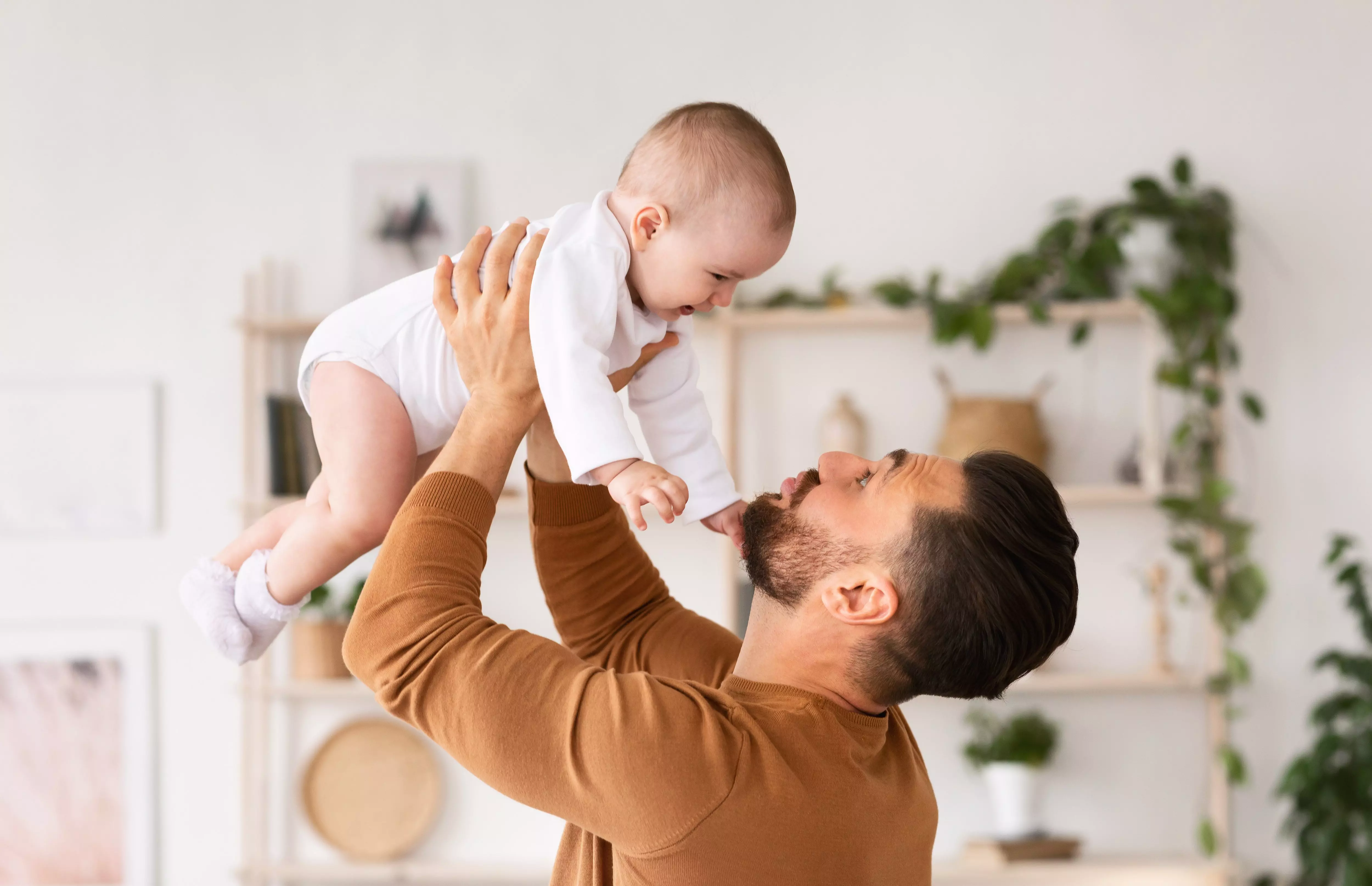 Father lifting baby wearing white Pepco bodysuit playfully in the air at home.