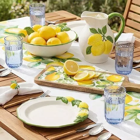 Stół ogrodowy z zastawą w cytrynowy wzór, misą cytryn, dzbankiem i niebieskimi szklankami.