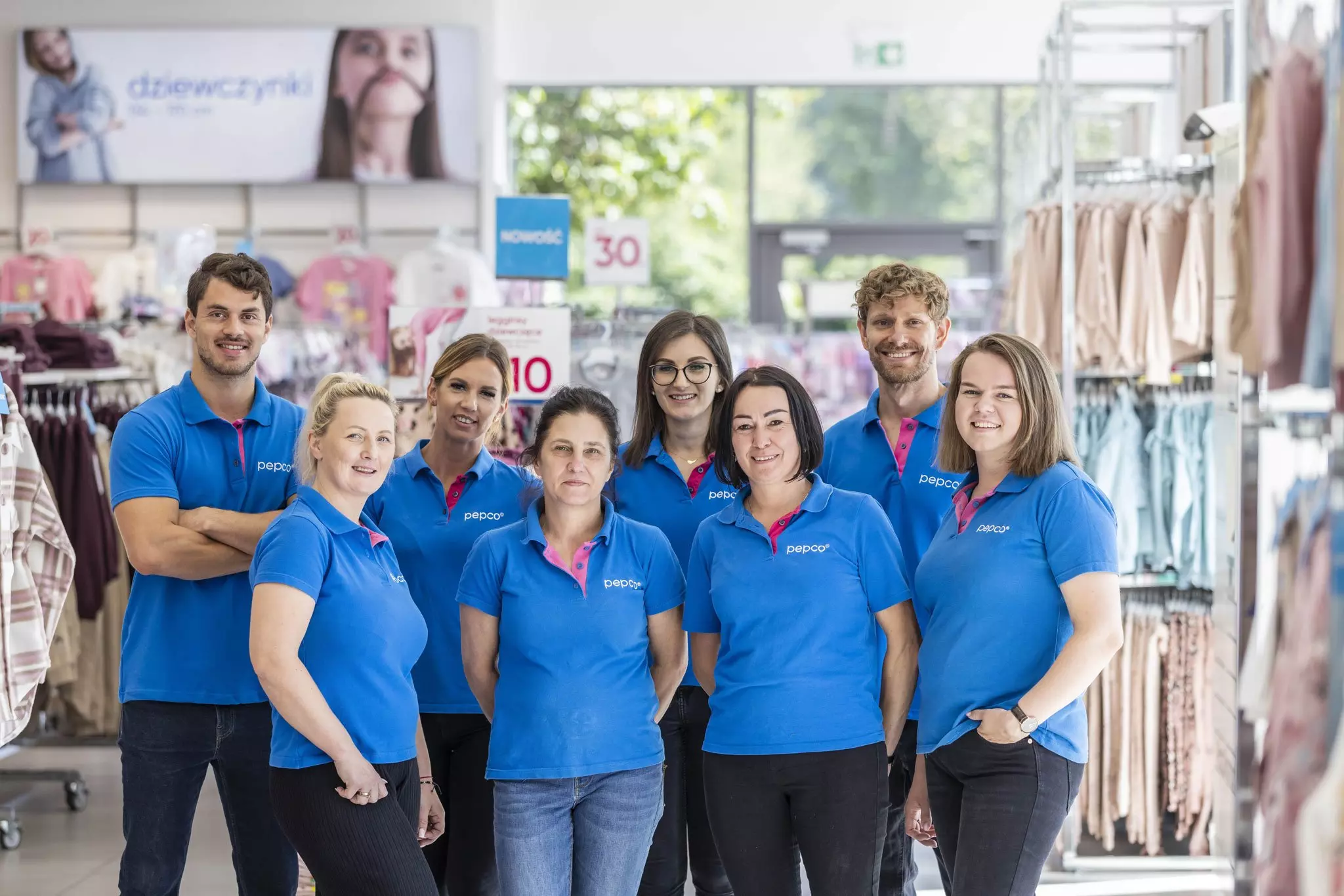 Pepco store staff wearing blue polo shirts with pink accents standing together in a bright retail space with clothing displays.