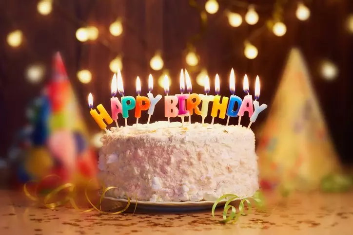 Pepco birthday cake with lit candles spelling Happy Birthday, surrounded by party hats and decorative lights.