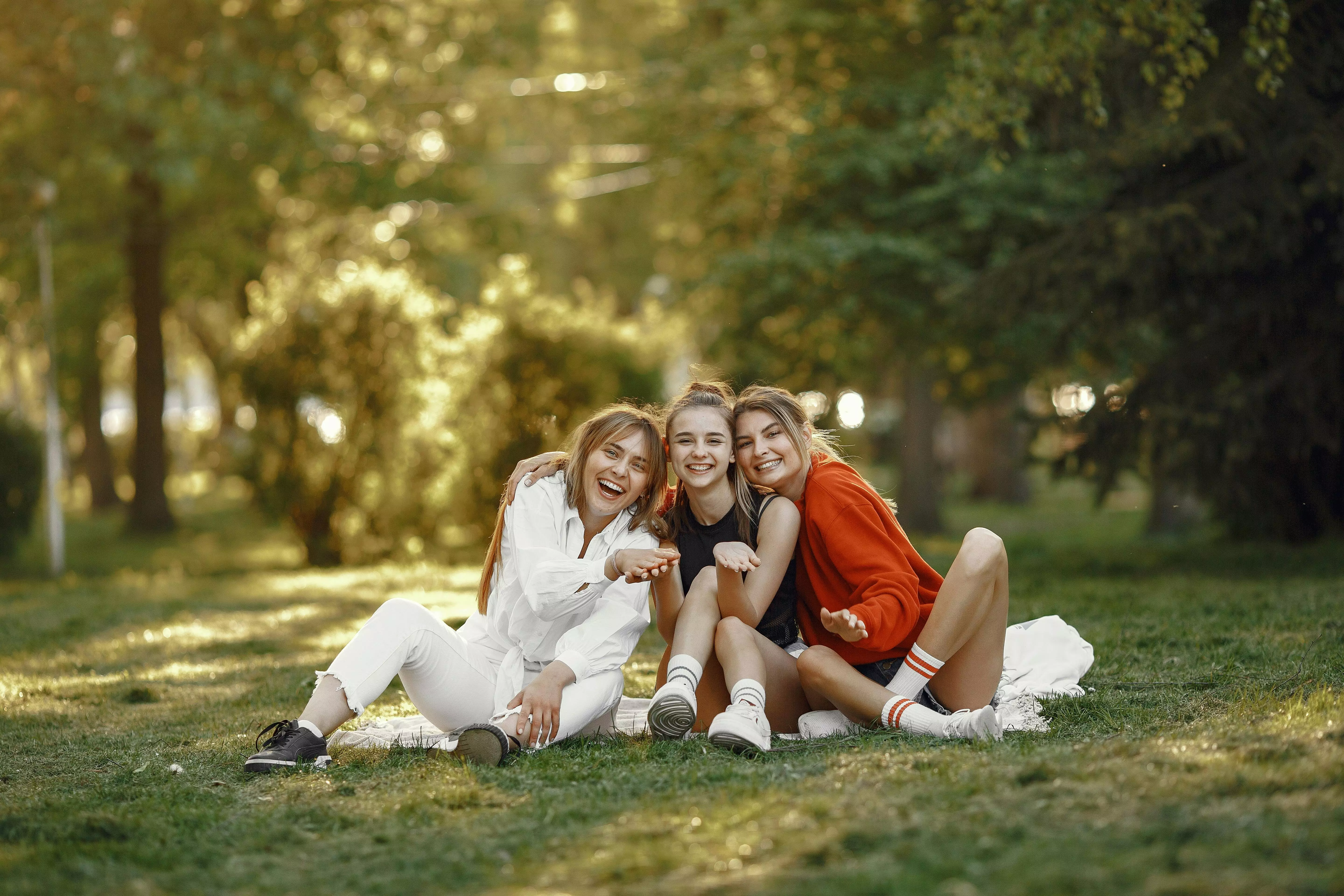 Tre ragazze sorridenti sedute sull'erba in un parco soleggiato indossando abbigliamento casual Pepco.