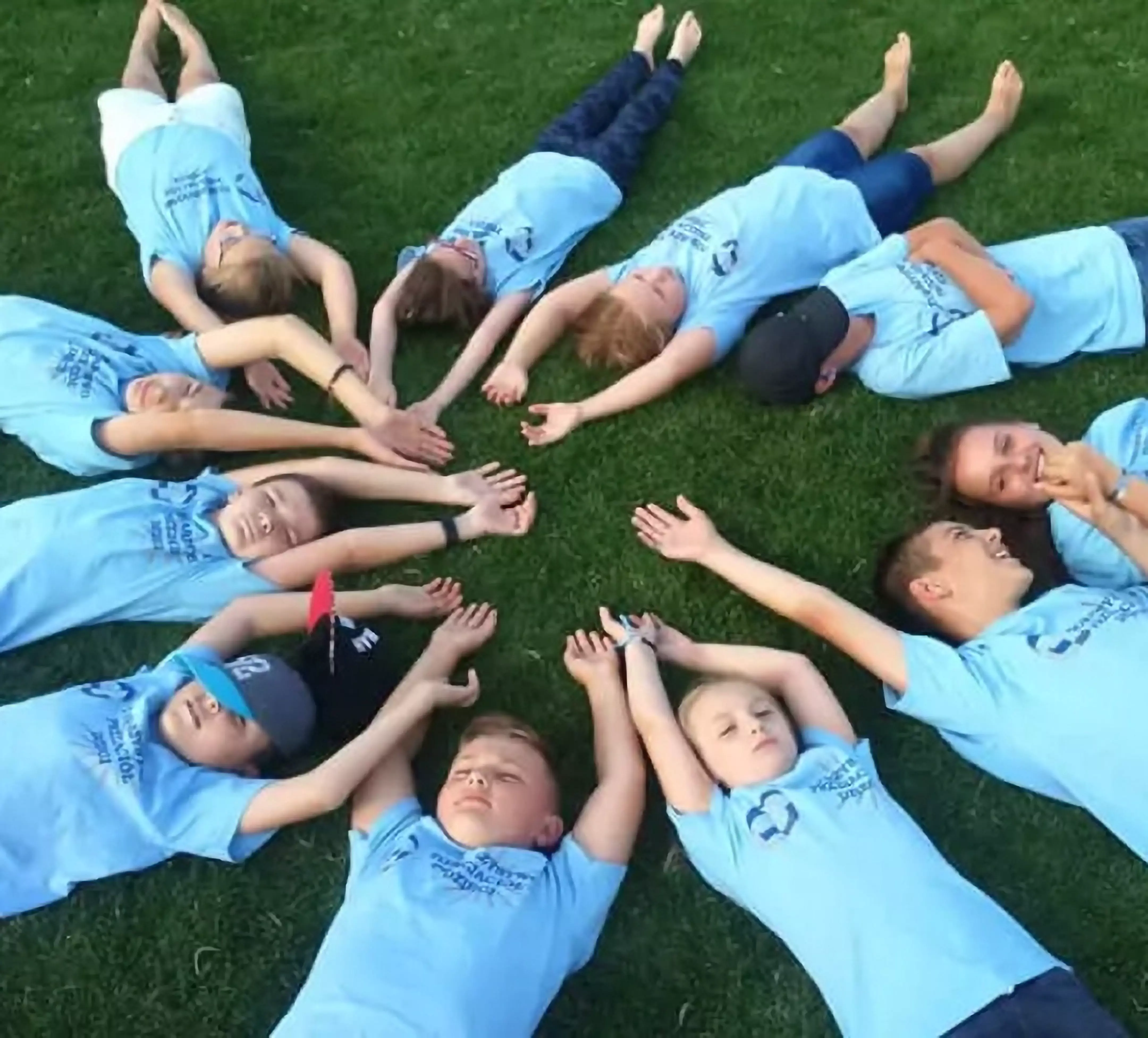 Children lying in a circle on grass wearing light blue Pepco t-shirts, reaching their hands toward the center.