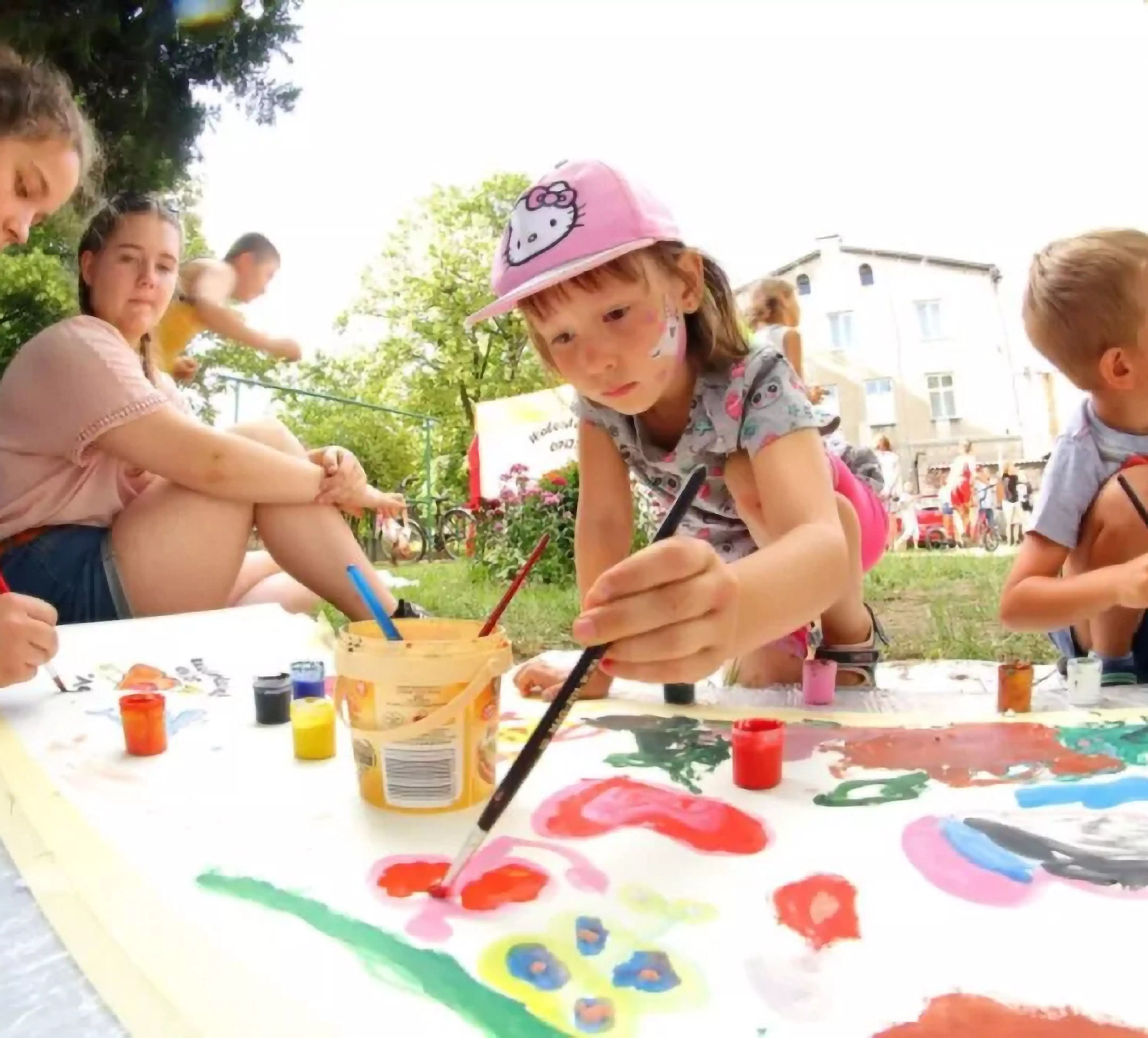 Child in pink Hello Kitty cap painting outdoors with colorful paints from Pepco, with other children around the table.