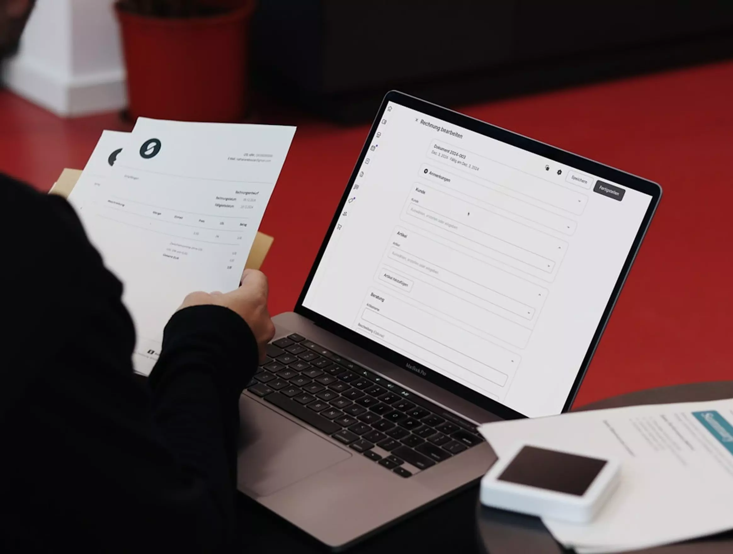 Person reviewing invoice document while using laptop displaying billing software on red desk.