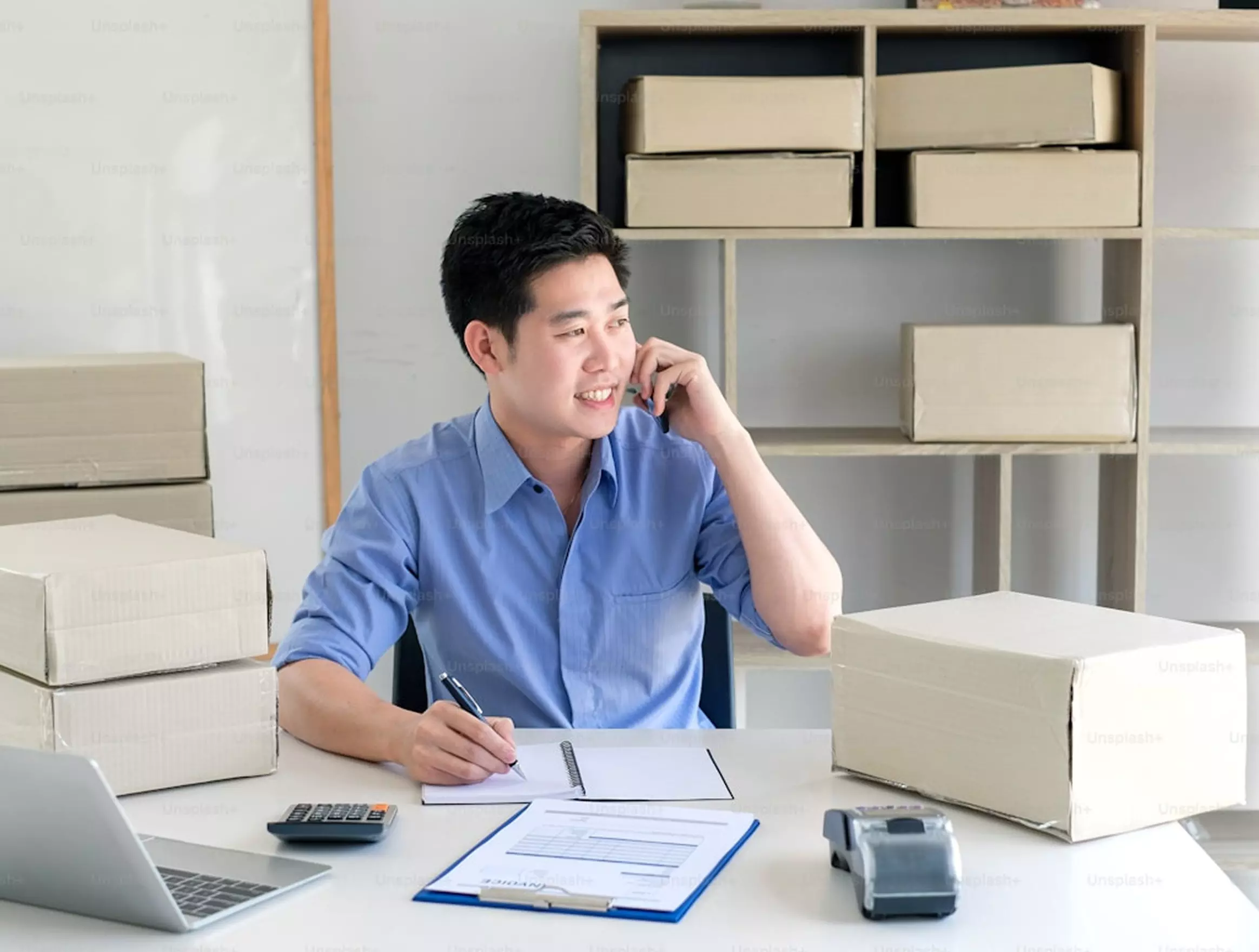 Man in blue shirt talking on phone while taking notes, surrounded by shipping boxes in workspace.