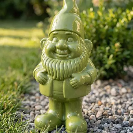 Zielona ceramiczna figurka krasnala ogrodowego z brodą i spiczastą czapką stojąca na żwirze.