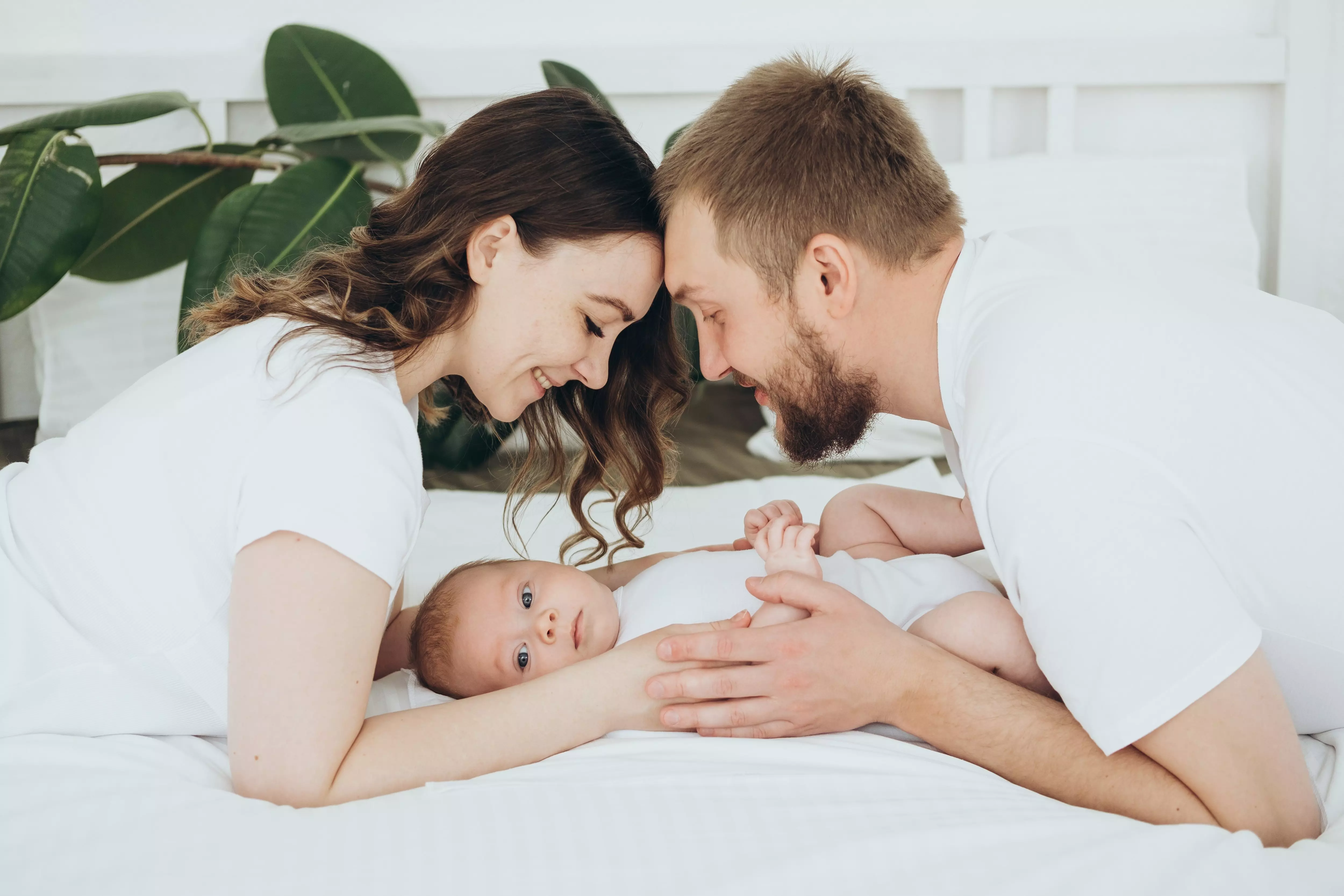 Pepco parents smiling at their baby lying on white bedding in a bright bedroom.