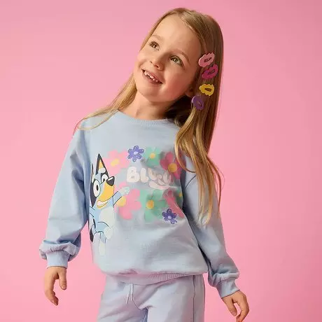 Dziewczynka w jasnoniebieskiej bluzie z kolorowymi kwiatami i postaciami, z ozdobnymi spinkami we włosach.