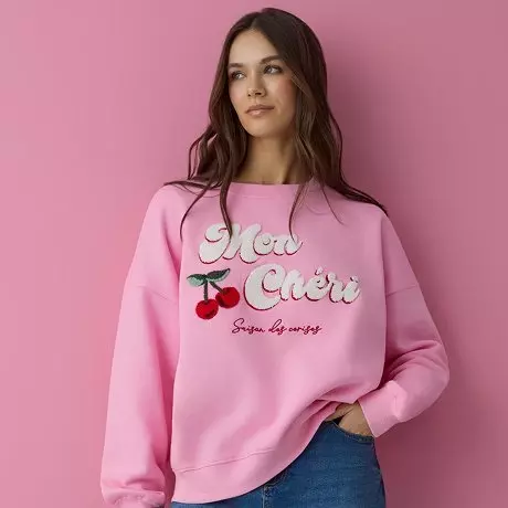Różowa bluza z napisem "Mon Chéri" i motywem wiśni, noszona przez osobę na różowym tle.
