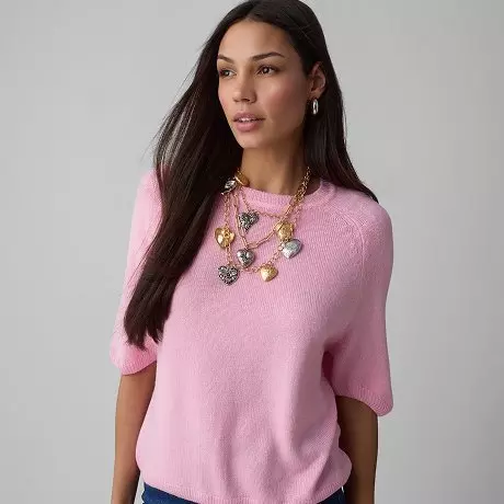 Kobieta w różowym swetrze z krótkim rękawem od Pepco, ozdobiona złotym naszyjnikiem z ozdobnymi zawieszkami.