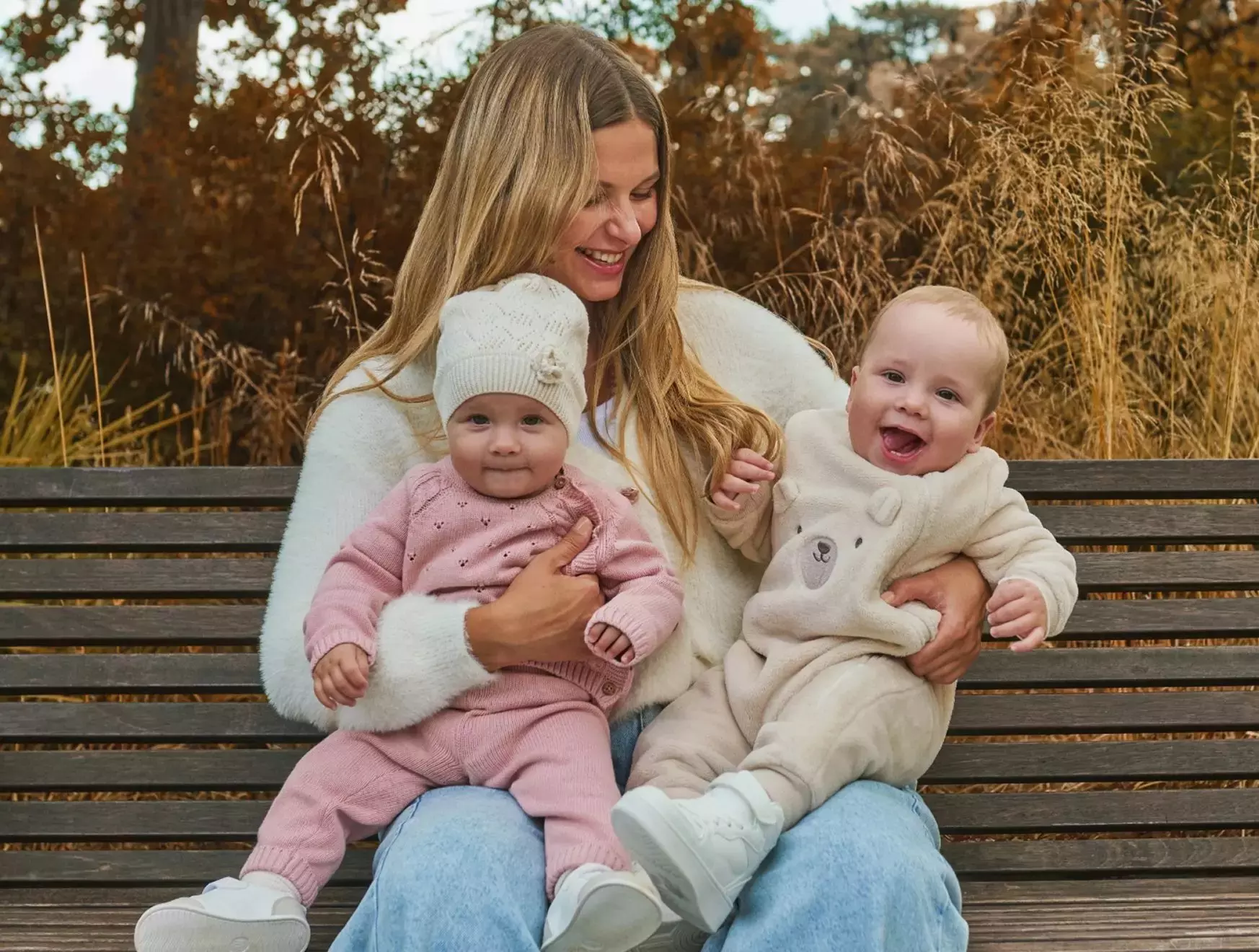 Pepco mother sitting on park bench holding two babies wearing pink and cream knitwear outdoors in autumn.