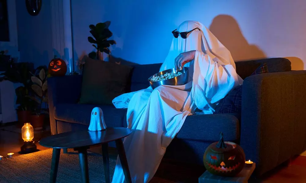 Osoba w przebraniu ducha z Pepco, siedząca na kanapie z miską przekąsek, otoczona dekoracjami halloweenowymi i dyniami.