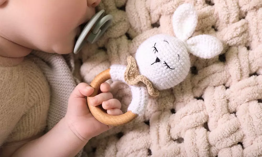 Bebé sosteniendo un sonajero de oveja de peluche con aro de madera sobre manta texturizada.