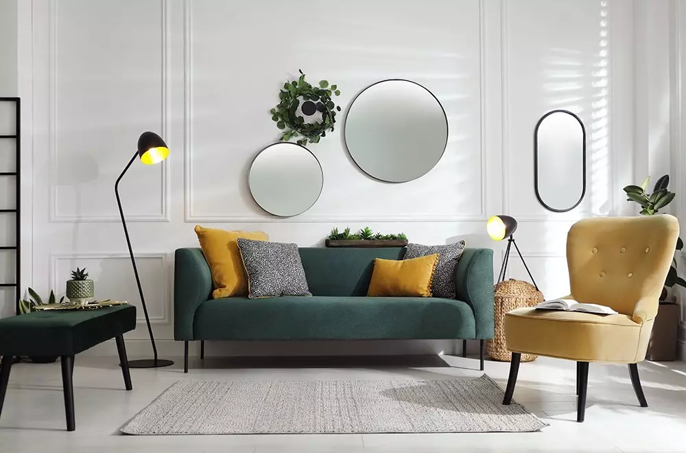 Elegancki salon Pepco z zieloną sofą, żółtym fotelem, okrągłymi lustrami i dekoracyjnymi lampami na białej ścianie.