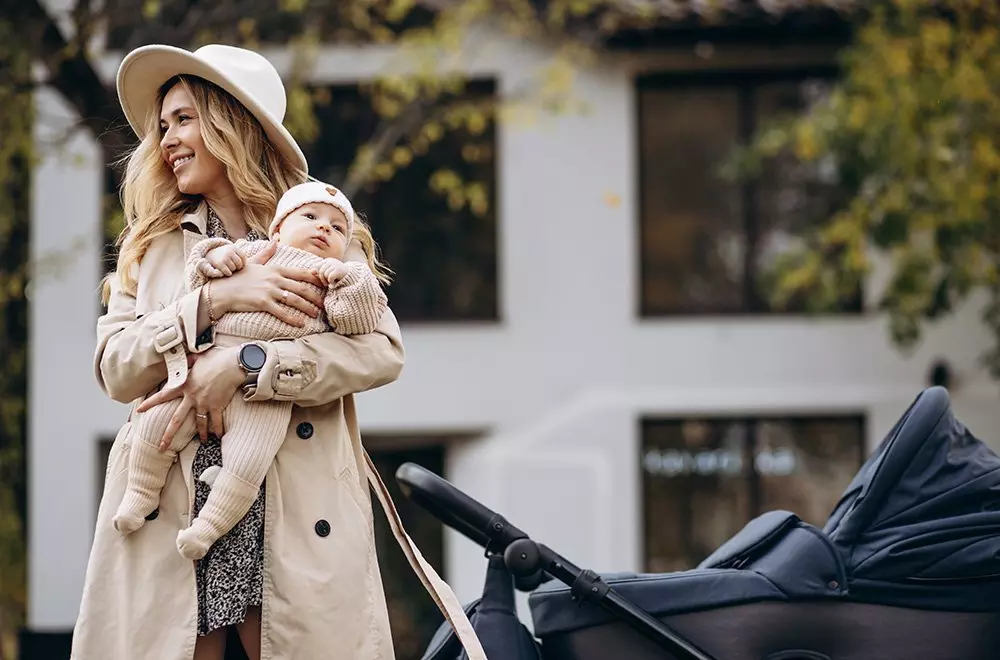 Osoba w beżowym płaszczu od Pepco trzymająca niemowlę w dzianinowym ubranku, obok widoczny wózek dziecięcy.