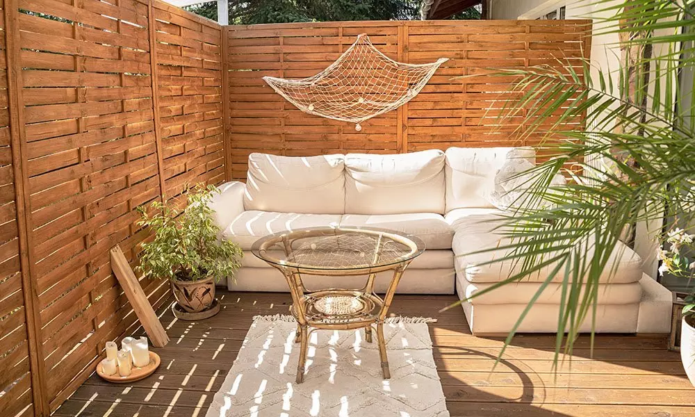 Przytulny kącik wypoczynkowy z białą sofą narożną, szklanym stolikiem i dekoracjami ogrodowymi Pepco na drewnianym tarasie.