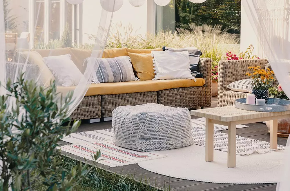 Przytulny taras z wiklinową sofą Pepco, poduszkami dekoracyjnymi, pufem i stolikiem kawowym w otoczeniu roślin.