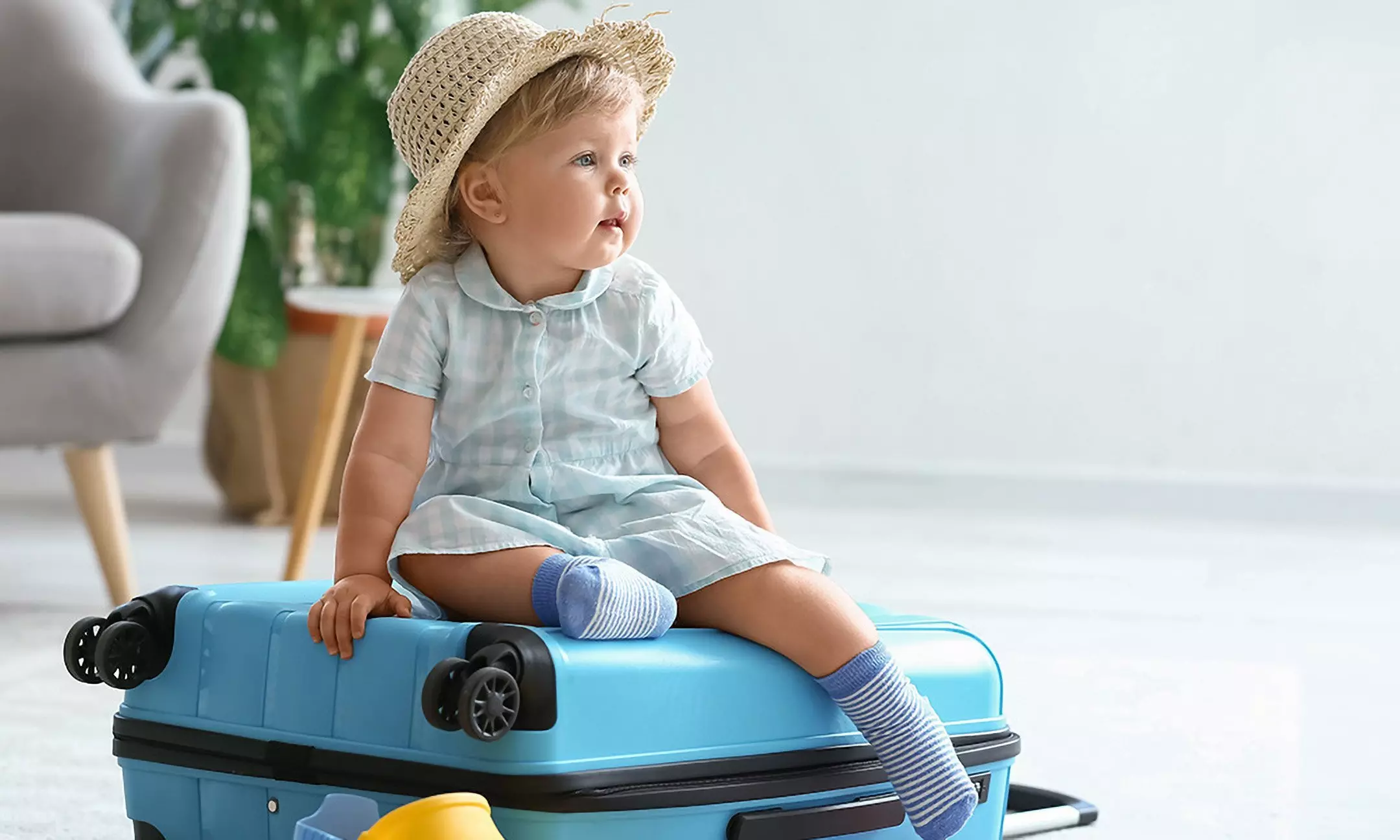 Dziecko w słomkowym kapeluszu i błękitnym komplecie Pepco siedzi na niebieskiej walizce, ubrane w pasiaste skarpetki.