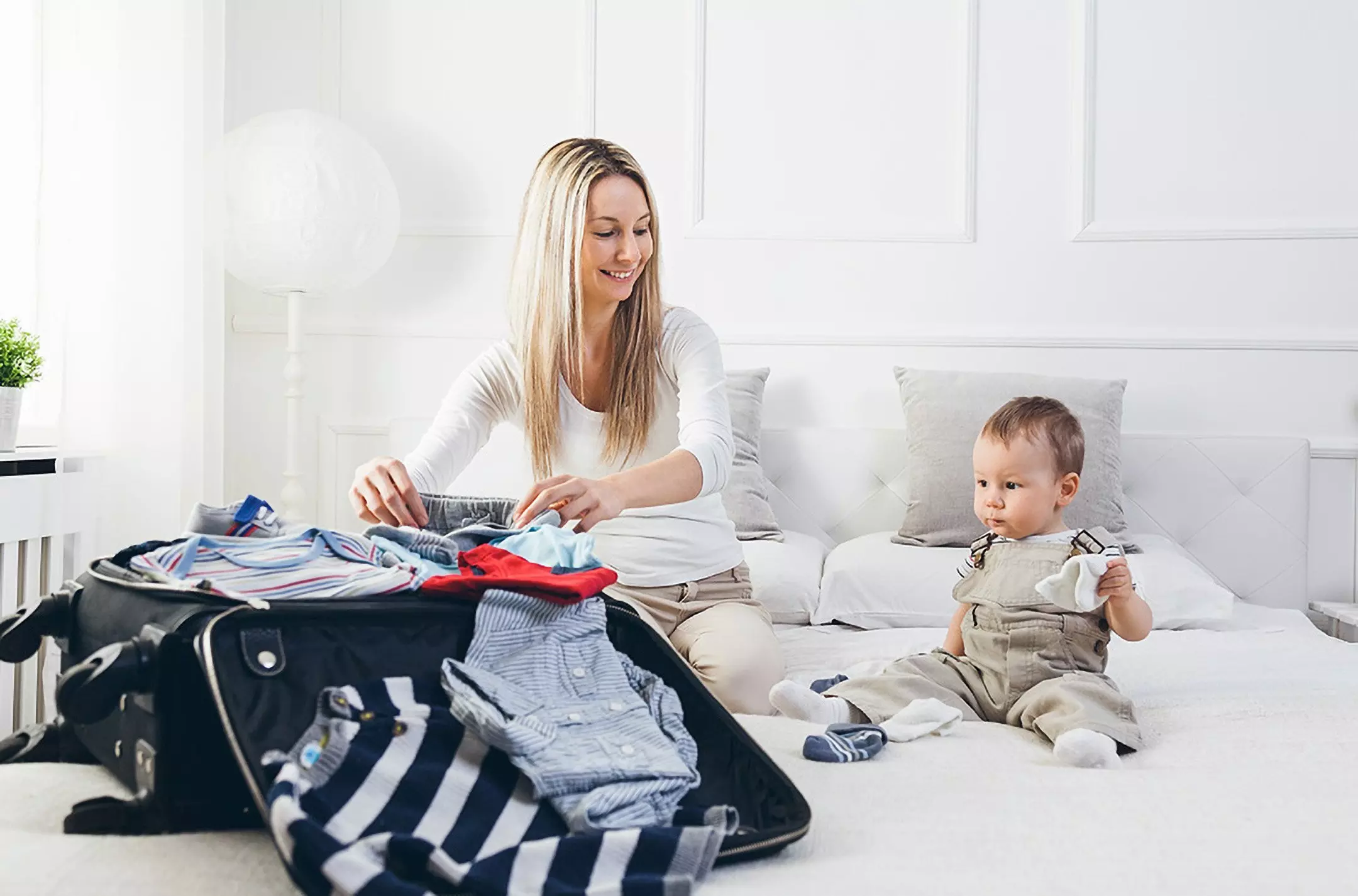 Woman packing Pepco children's clothing into suitcase with toddler sitting nearby on white sofa.