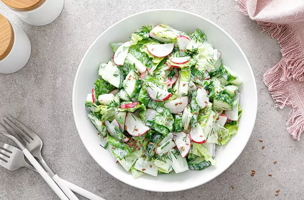 Fresh salad with radishes and greens in a Pepco white bowl, displayed on a gray surface with forks and pink napkin.