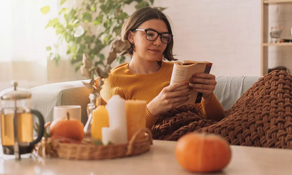 Osoba w żółtym swetrze Pepco czyta książkę otulona brązowym kocem, obok koszyk z dynią, świecami i sokiem.