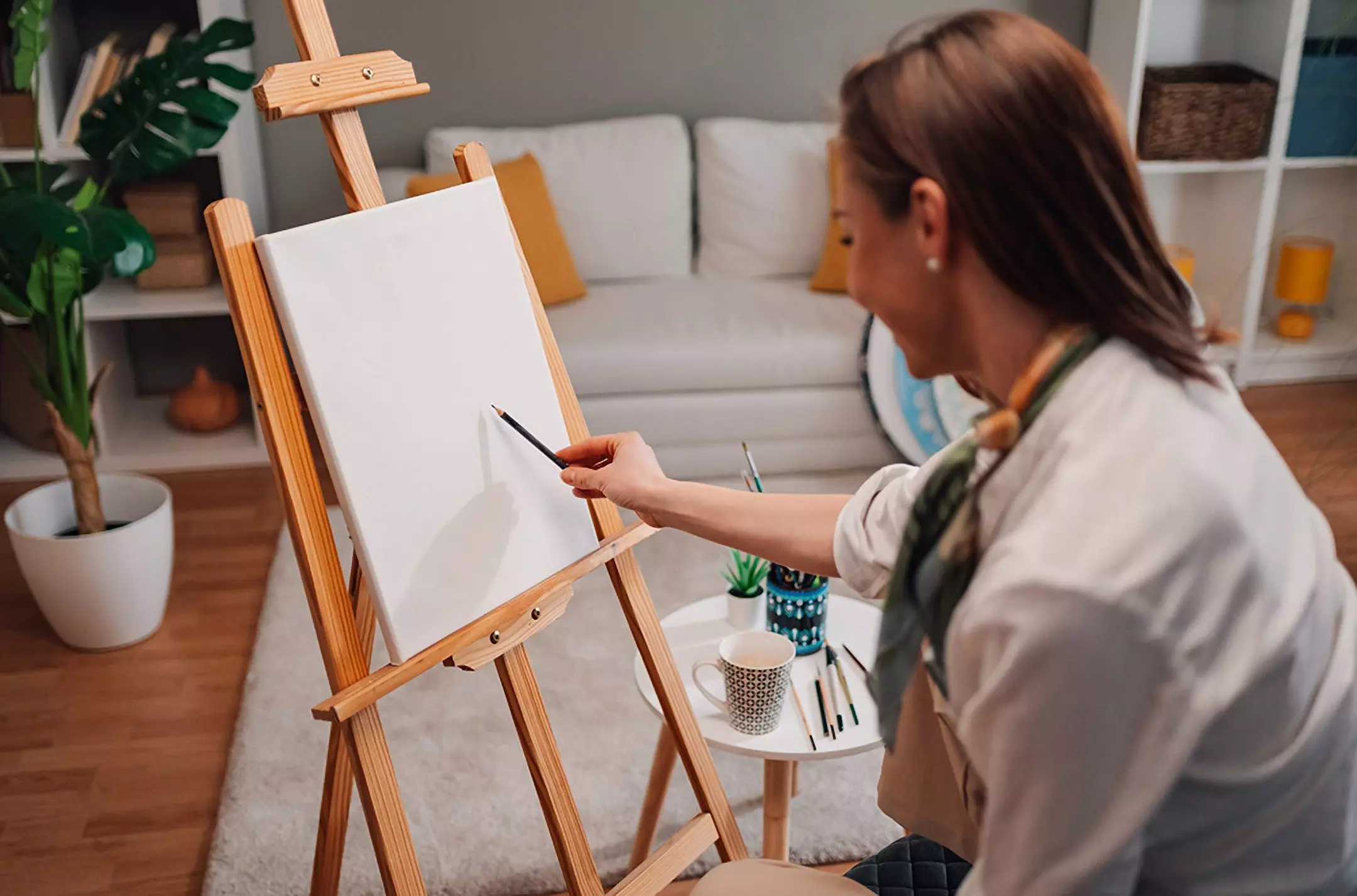 Kobieta maluje na drewnianej sztaludze w przytulnym salonie, obok akcesoria malarskie Pepco na małym stoliku.