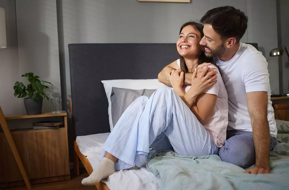 Para siedząca na łóżku w przytulnej sypialni, ubrana w wygodną odzież Pepco, dzieli radosny moment.