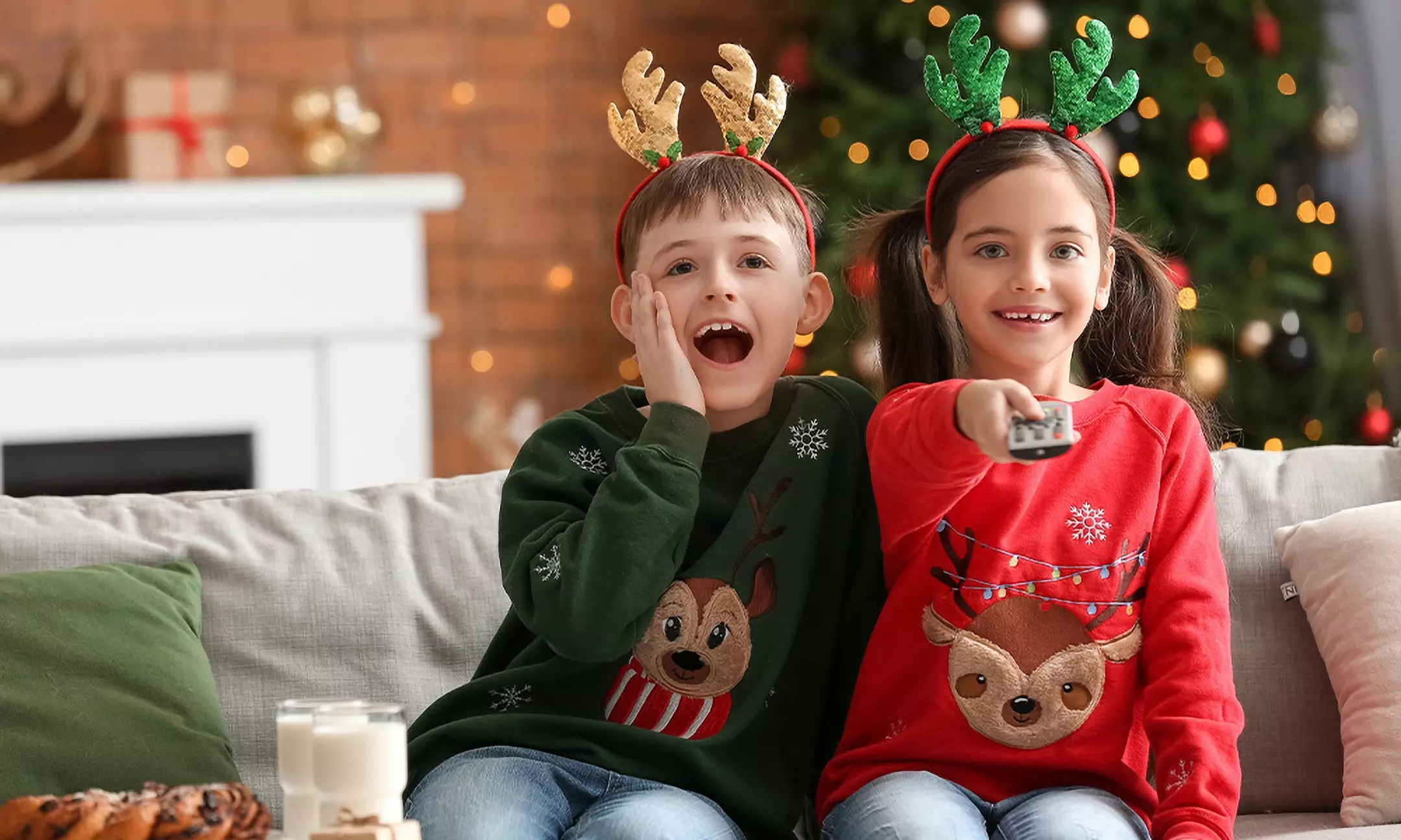 Gyerekek Pepco karácsonyi pulóverekben és rénszarvas fejdíszekkel a kanapén ülve, háttérben karácsonyfával.