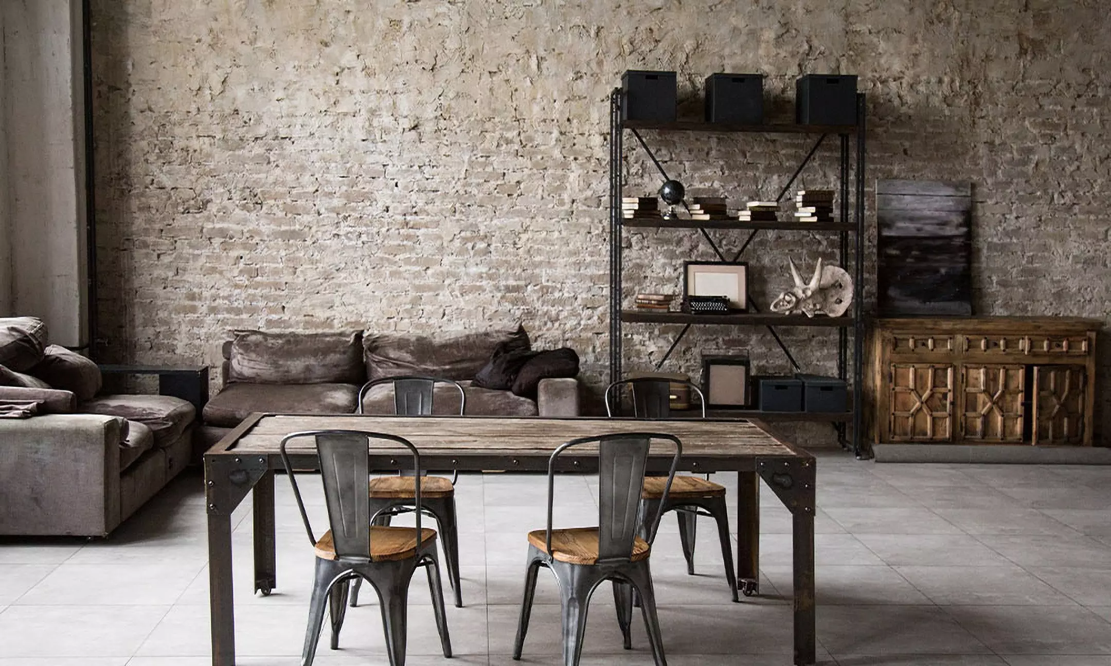 Industrialne wnętrze z meblami Pepco - stół z metalowymi krzesłami, sofa i regał z dekoracjami na tle ceglanej ściany.