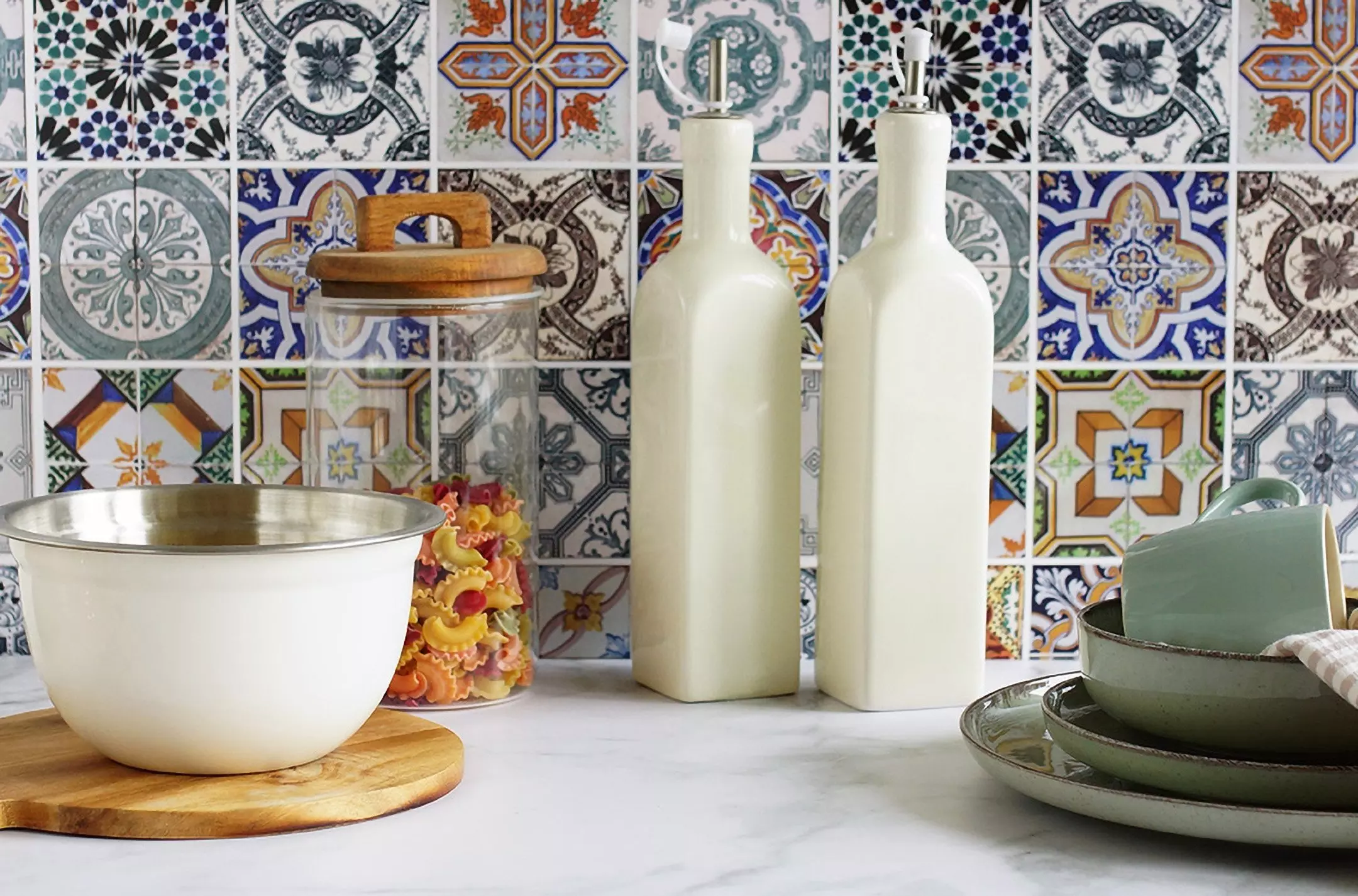 Kolekcja kuchenna Pepco z białymi butelkami, miską, słoikiem z drewnianą pokrywką i zielonymi talerzami na tle kolorowych kafelków.