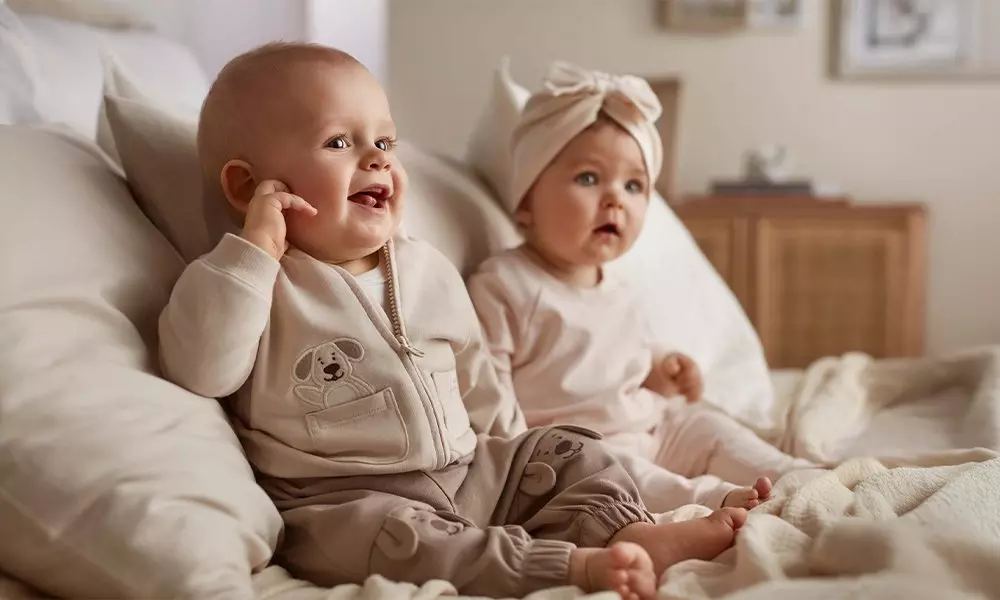 Two babies sitting on a bed wearing soft beige outfits, one with a dog design and one with a bow headband.