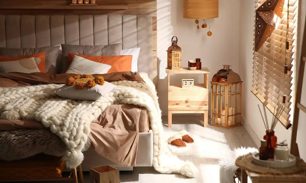 Dormitor confortabil Pepco cu pat tapițat, pături pufoase, perne portocalii și decorațiuni calde, inclusiv felinare și difuzor de aromă.