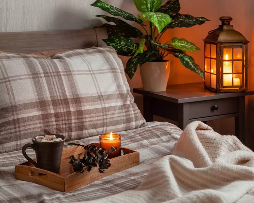 Przytulna sypialnia Pepco z kratką pościelą, drewnianą tacą z kubkiem i świecą, rośliną doniczkową i latarnią na szafce nocnej.