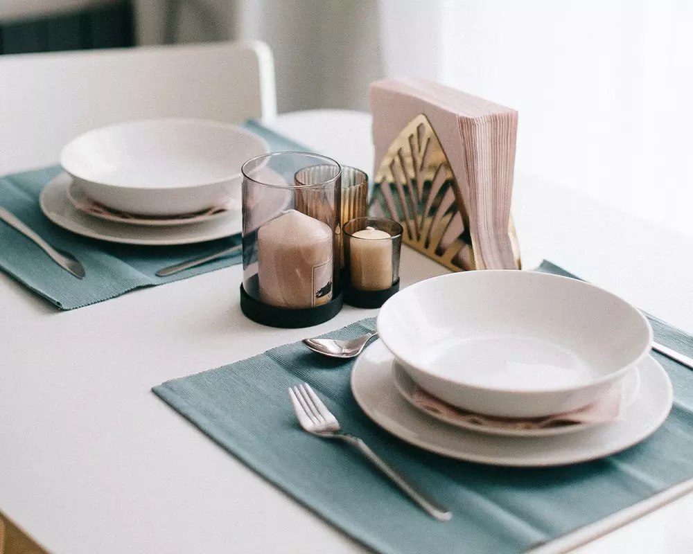 Elegancka zastawa stołowa Pepco z białymi talerzami, świeczkami i serwetnikiem na niebieskich podkładkach.