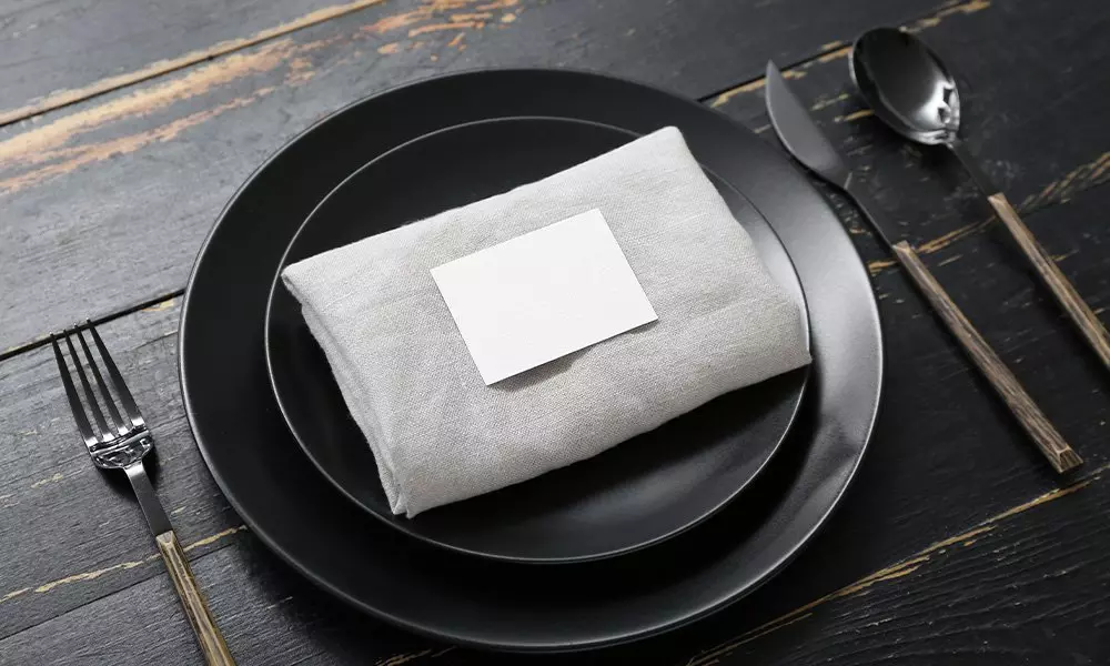 Eleganckie czarne talerze Pepco z lnianą serwetką i pustą wizytówką, obok sztućce na drewnianym stole.