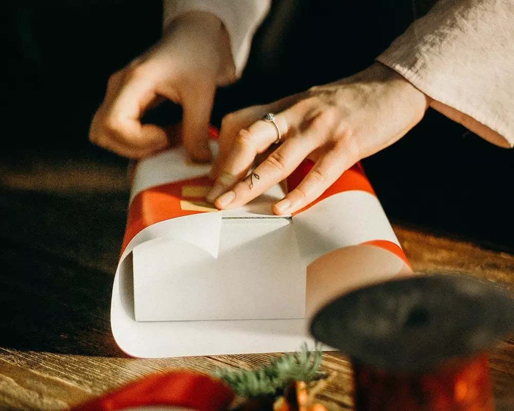 Hands wrapping a Pepco gift in orange and white paper on a wooden table with festive decorations visible.