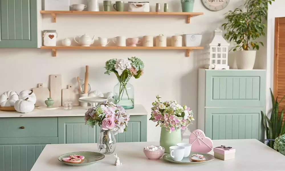 Pepco otthoni dekorációs termékek pasztell zöld konyhában, virágcsokrokkal, polcokon teáskészletekkel és dekoratív tárgyakkal.