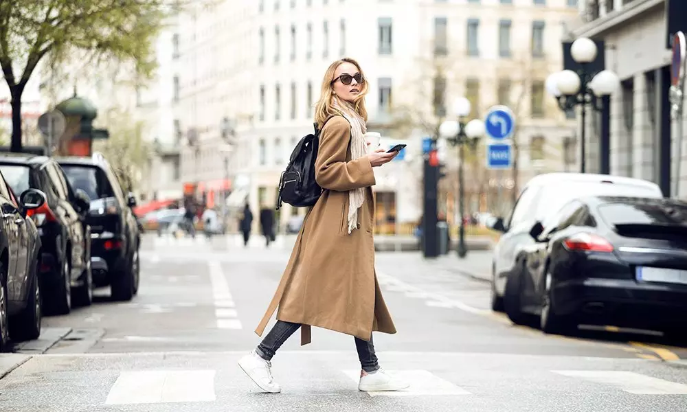 Woman wearing Pepco camel coat with white scarf and backpack, checking phone while crossing a city street.