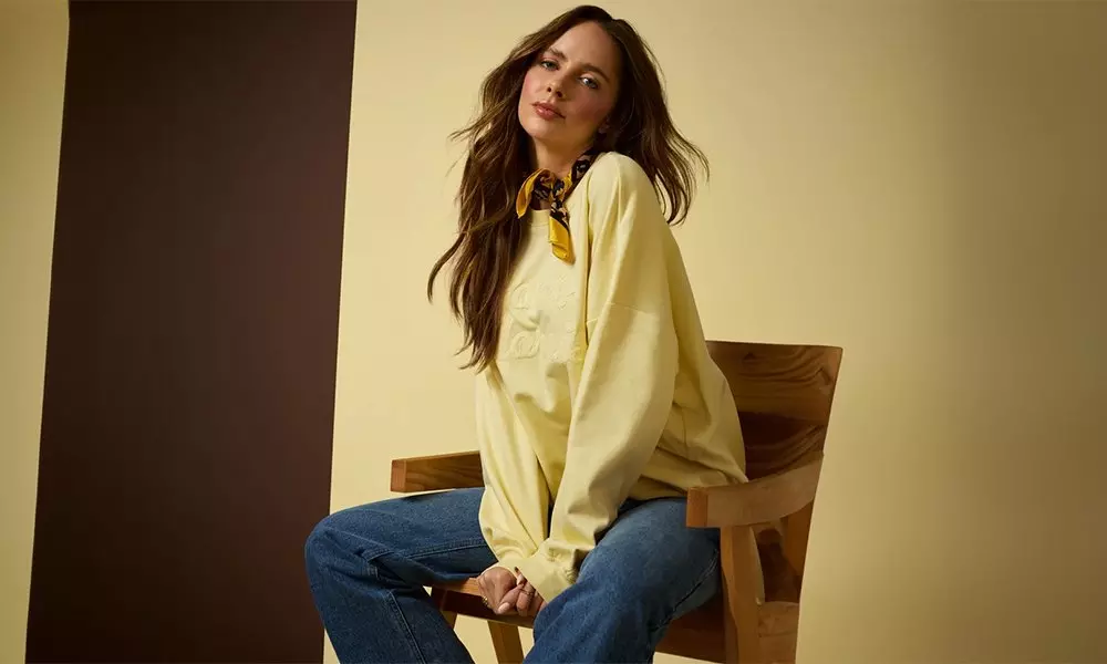 Woman sitting on wooden chair wearing yellow shirt, blue jeans, and patterned neck scarf.