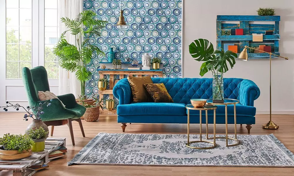 Pepco elegancki salon z niebieską pikowaną sofą, zielonym fotelem i roślinami, z wzorzystą tapetą i złotymi akcentami.