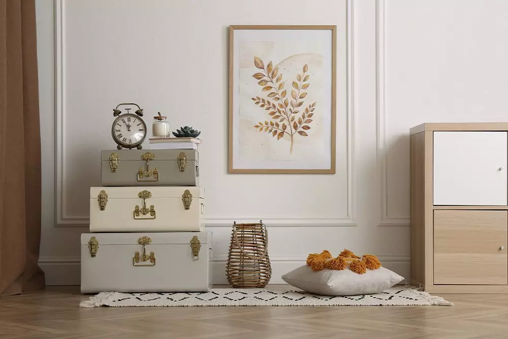 Pepco home decor featuring stacked vintage suitcases, botanical wall art, alarm clock, decorative pillow with tassels and wicker lantern on patterned rug.