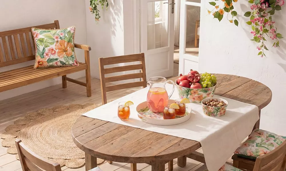 Wooden outdoor dining table set with iced drinks, fresh fruit, and nuts on a sunny patio.