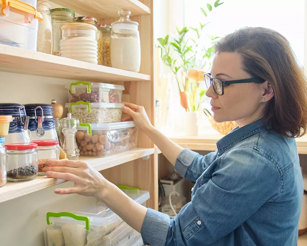 Kobieta układająca pojemniki z Pepco w drewnianej spiżarni, przechowująca produkty spożywcze w szklanych słoikach i plastikowych pudełkach.