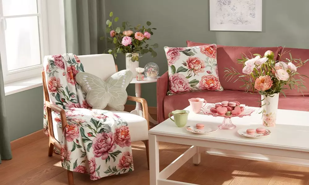 Cozy living room with pink floral decor featuring armchair with throw blanket, coral sofa, and table with macarons and fresh flowers.