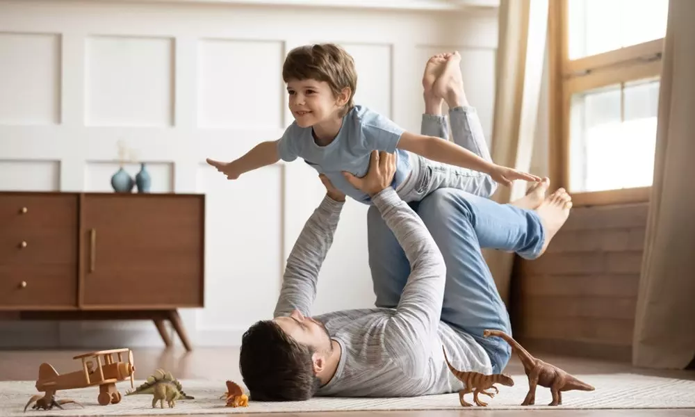 Dziecko w niebieskiej koszulce Pepco udające samolot, uniesione przez dorosłego leżącego na dywanie, obok zabawkowe dinozaury.