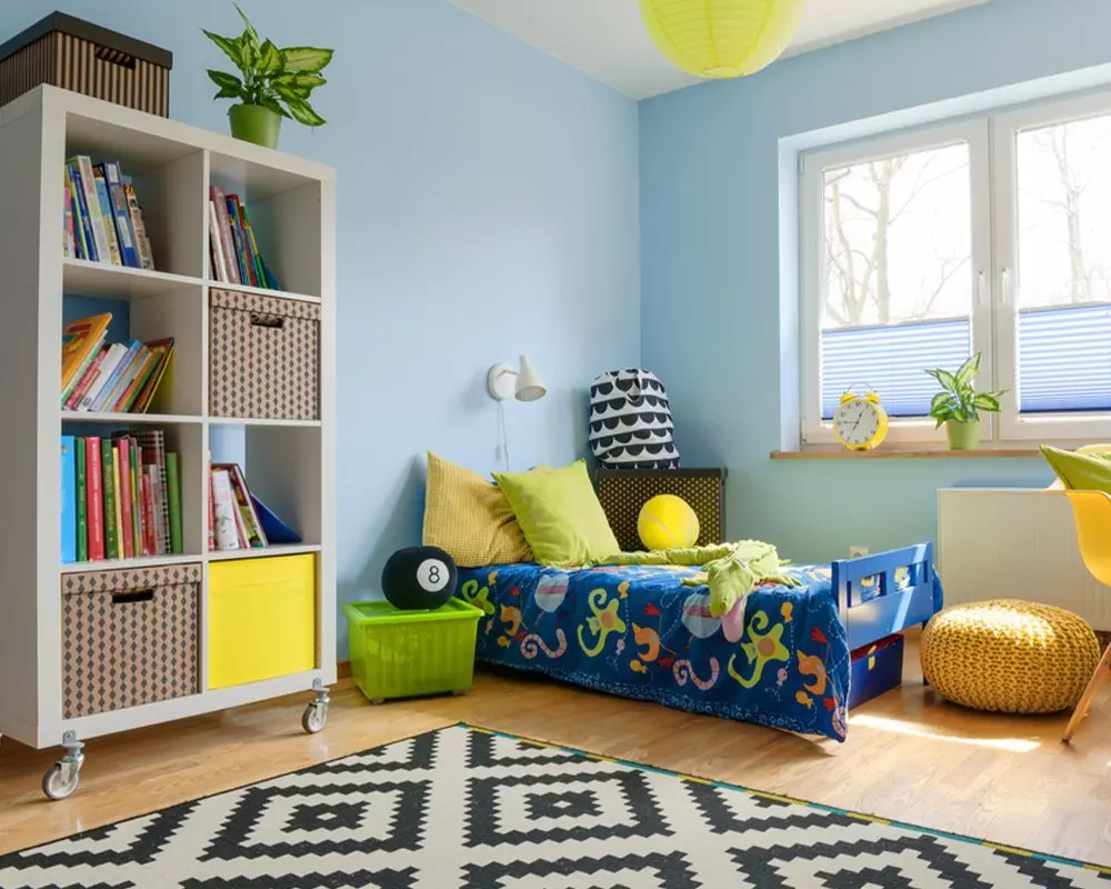 Jasny pokój dziecięcy z meblami Pepco: niebieskie łóżko, biały regał na książki, kolorowe poduszki i dywan w geometryczne wzory.