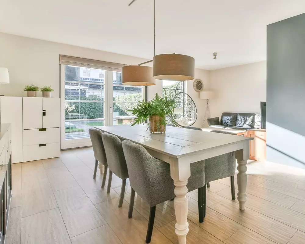 Nowoczesna jadalnia z meblami Pepco, drewniany stół z szarymi krzesłami, wiszącymi lampami i roślinami w jasnym pomieszczeniu.