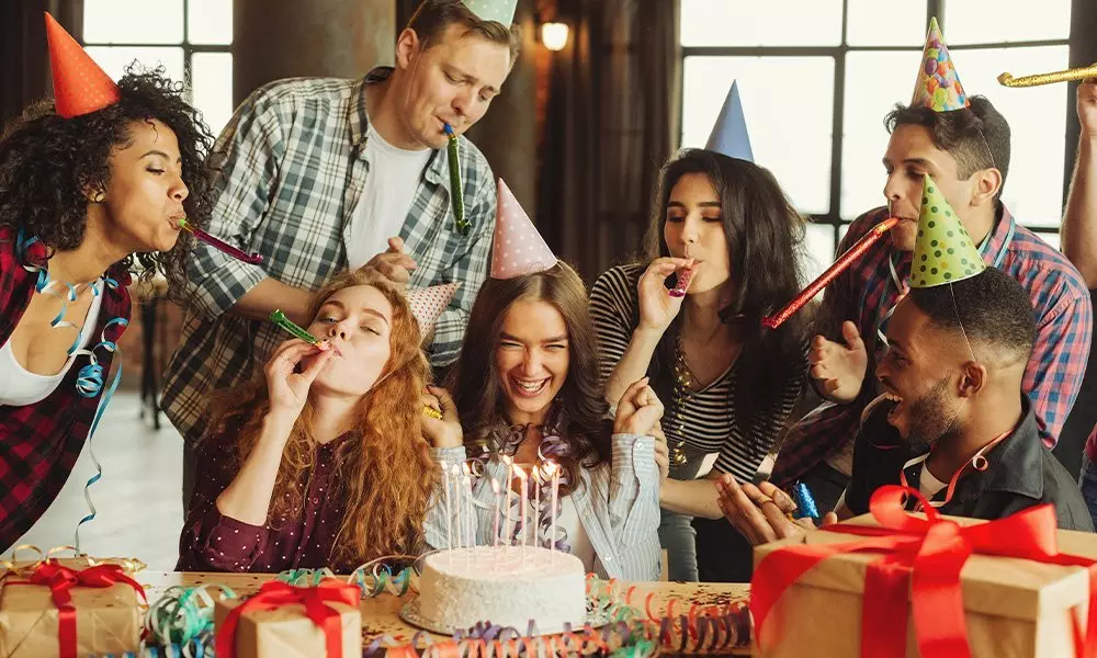 A birthday celebration with people wearing party hats, blowing noisemakers around a cake with candles. Pepco party supplies visible.