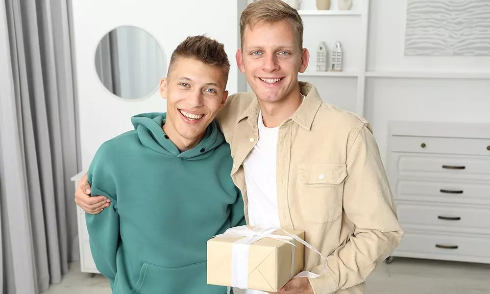 Dwóch mężczyzn w ubraniach Pepco, jeden w zielonej bluzie z kapturem, drugi w beżowej koszuli, trzymający zapakowany prezent.